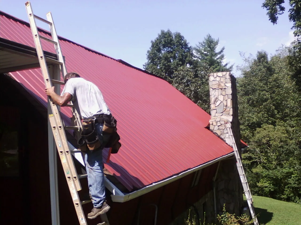 Roofer on a ladder beside a red metal roof and stone chimney — Soffit & Fascia Repair by Roofing Brothers in Hagerstown